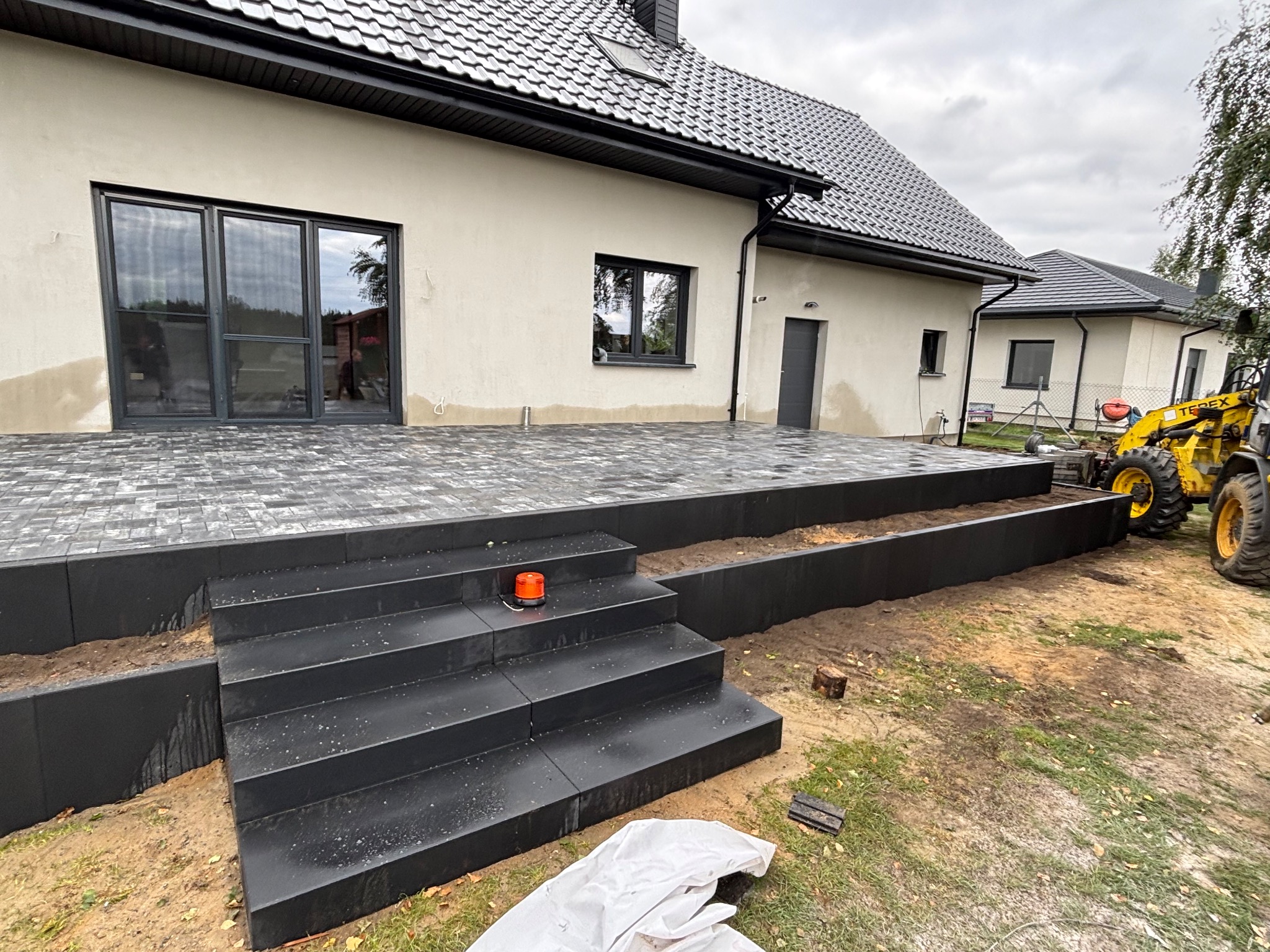 Nowoczesny taras z ciemnej kostki brukowej, schody z podświetleniem, jasny budynek w tle, częściowo widoczna maszyna budowlana. Minimalistyczny design.