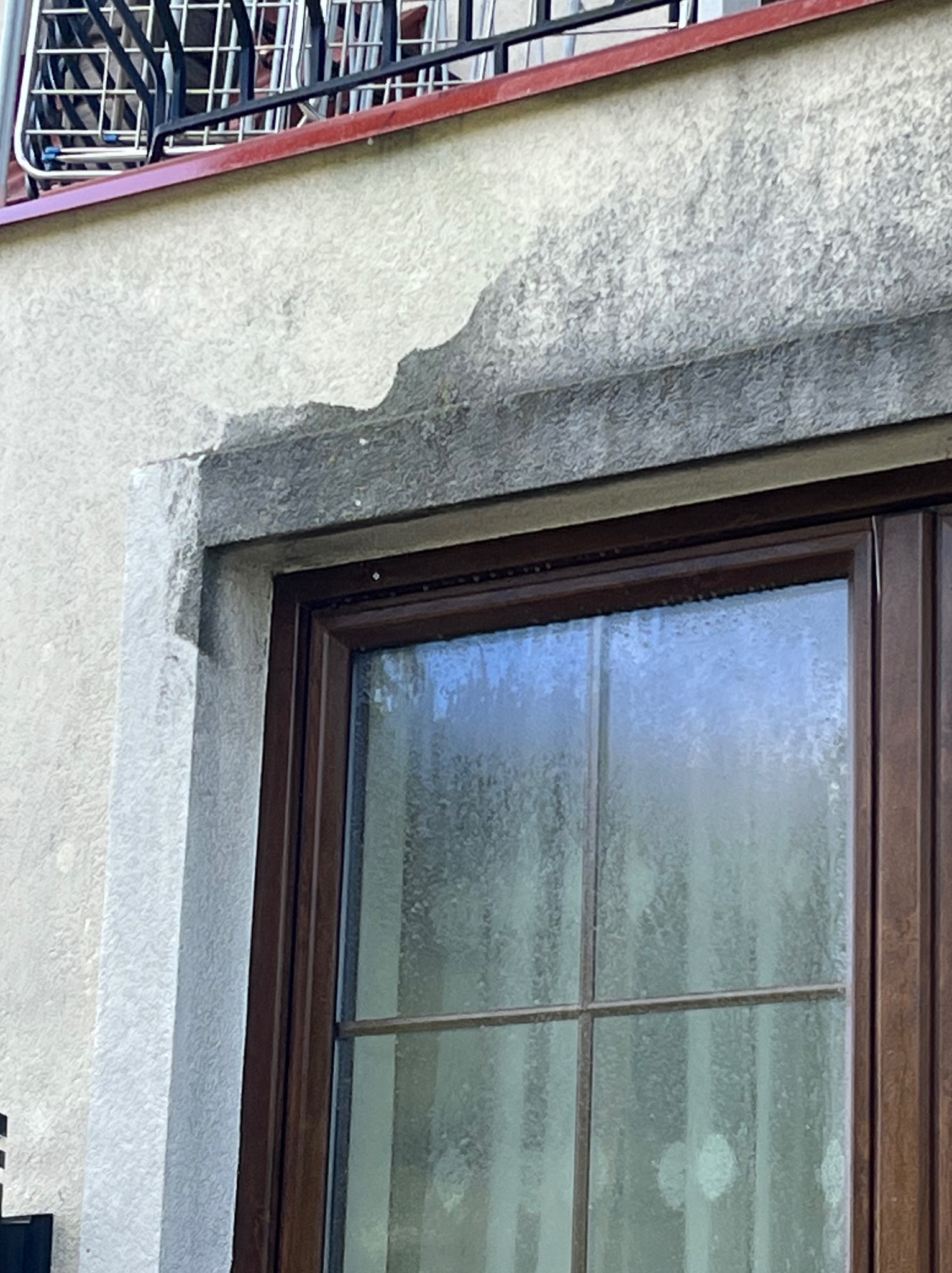 Fragment elewacji z widocznym uszkodzeniem tynku nad oknem z brązową ramą, na górze balkon z suszarką na pranie.