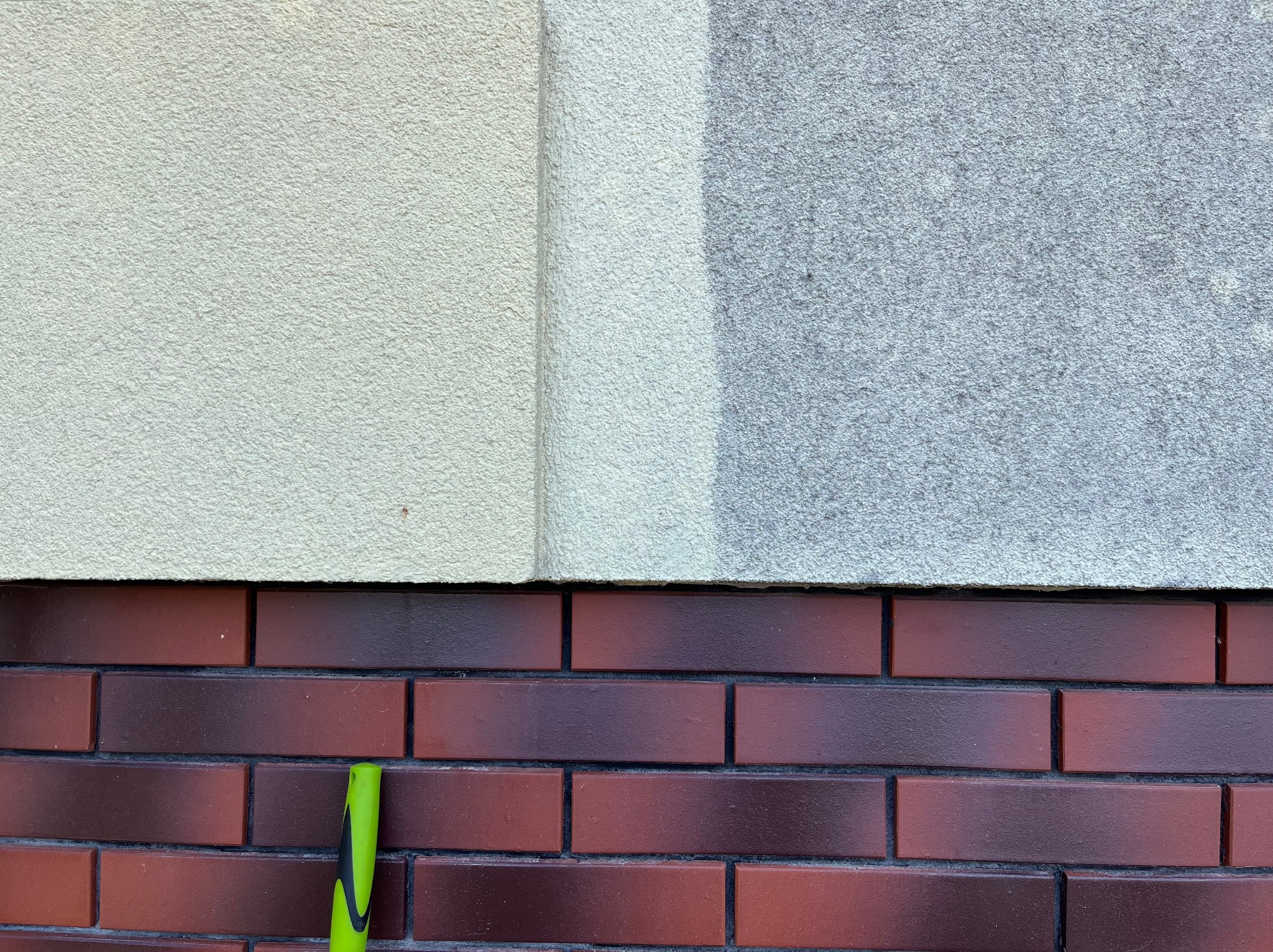 Fragment elewacji z cegły klinkierowej i jasnoszarego tynku strukturalnego; widoczny fragment zielonej rurki przy ścianie.