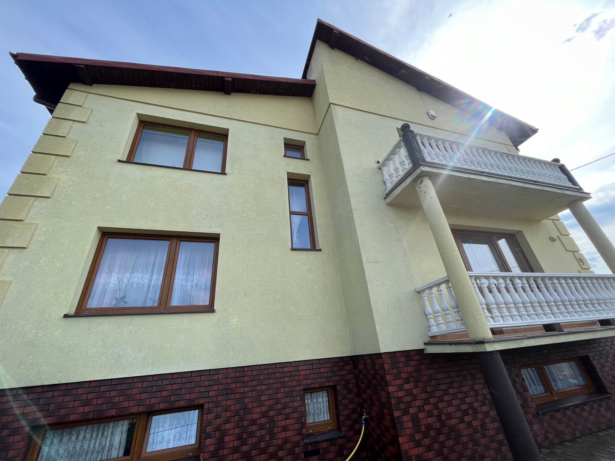 Widok z dołu na dwukondygnacyjny dom z kremową elewacją i ceglaną podmurówką, z balkonem z białą balustradą.