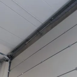 Wnętrze garażu z widoczną białą segmentową bramą garażową, szynami prowadzącymi i sprężynami naciągowymi, montowanymi pod sufitem z widocznymi elementami instalacji elektrycznej w korytkach.