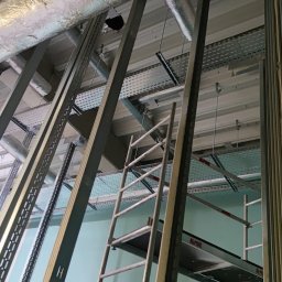 AMPERA POLSKA SPÓŁKA Z OGRANICZONĄ ODPOWIEDZIALNOŚCIĄ - Montaż metalowych korytek kablowych pod sufitem w hali przemysłowej, widoczne rury wentylacyjne i drabina rusztowania.