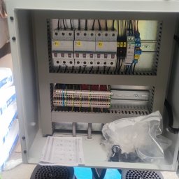 AMPERA POLSKA SPÓŁKA Z OGRANICZONĄ ODPOWIEDZIALNOŚCIĄ - Otwarta szafa sterownicza z widocznymi bezpiecznikami, listwami zaciskowymi i przewodami, częściowo zakryta przez dłoń trzymającą dokumentację techniczną, w tle worki z materiałami budowlanymi.