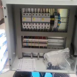 Otwarta szafa sterownicza z widocznymi bezpiecznikami, listwami zaciskowymi i przewodami, częściowo zakryta przez dłoń trzymającą dokumentację techniczną, w tle worki z materiałami budowlanymi.