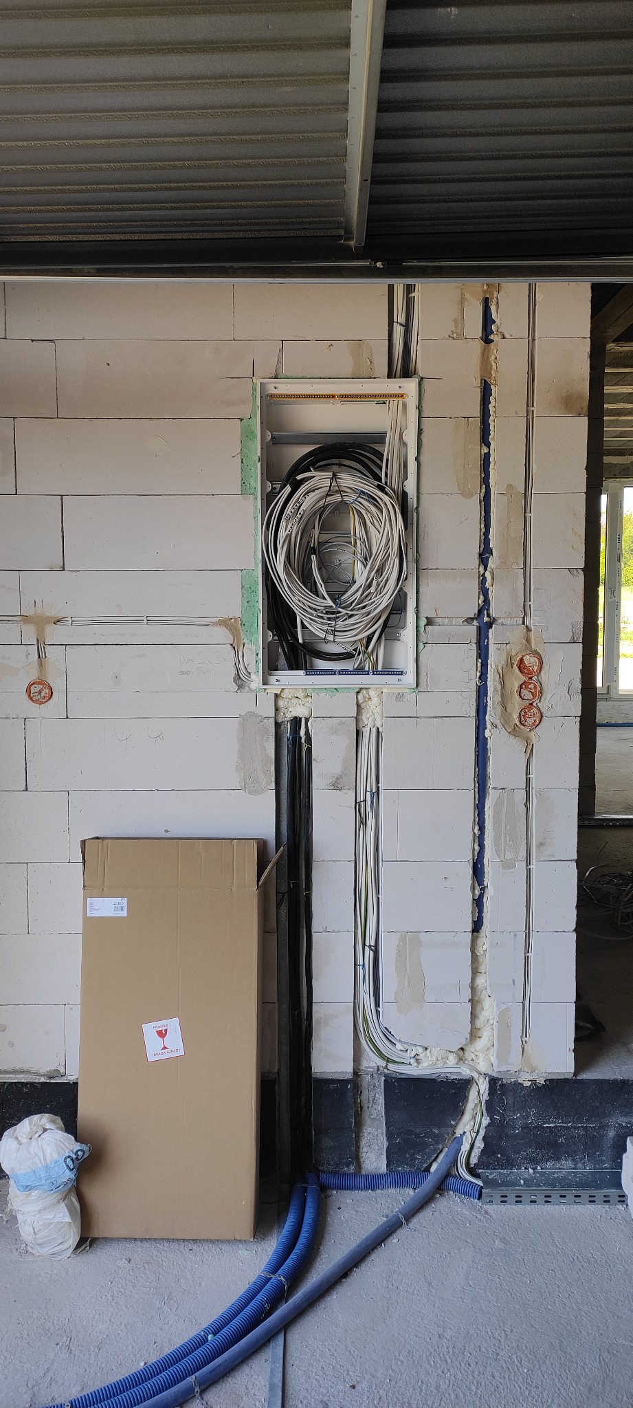 Wewnętrzna instalacja elektryczna w trakcie montażu w ścianie z betonu komórkowego, widoczne kable w puszce i rurki karbowane na podłodze, obok stoi kartonowe pudło z naklejką 'Fragile'.