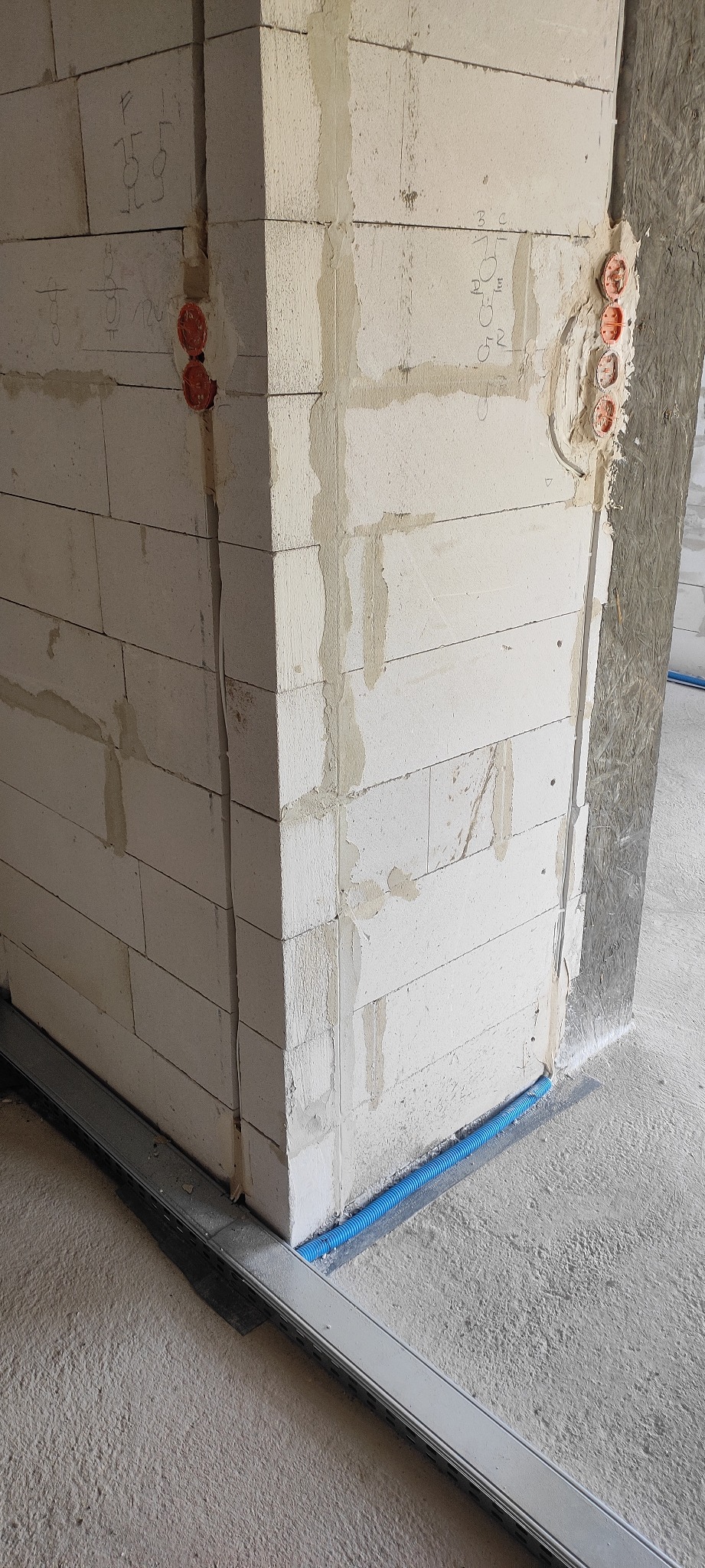 Murowany narożnik ściany z widocznymi puszkami elektrycznymi, bruzdami na kable i niebieską rurką karbowaną wzdłuż podłogi, na ścianie widoczne odręczne oznaczenia elektryczne.