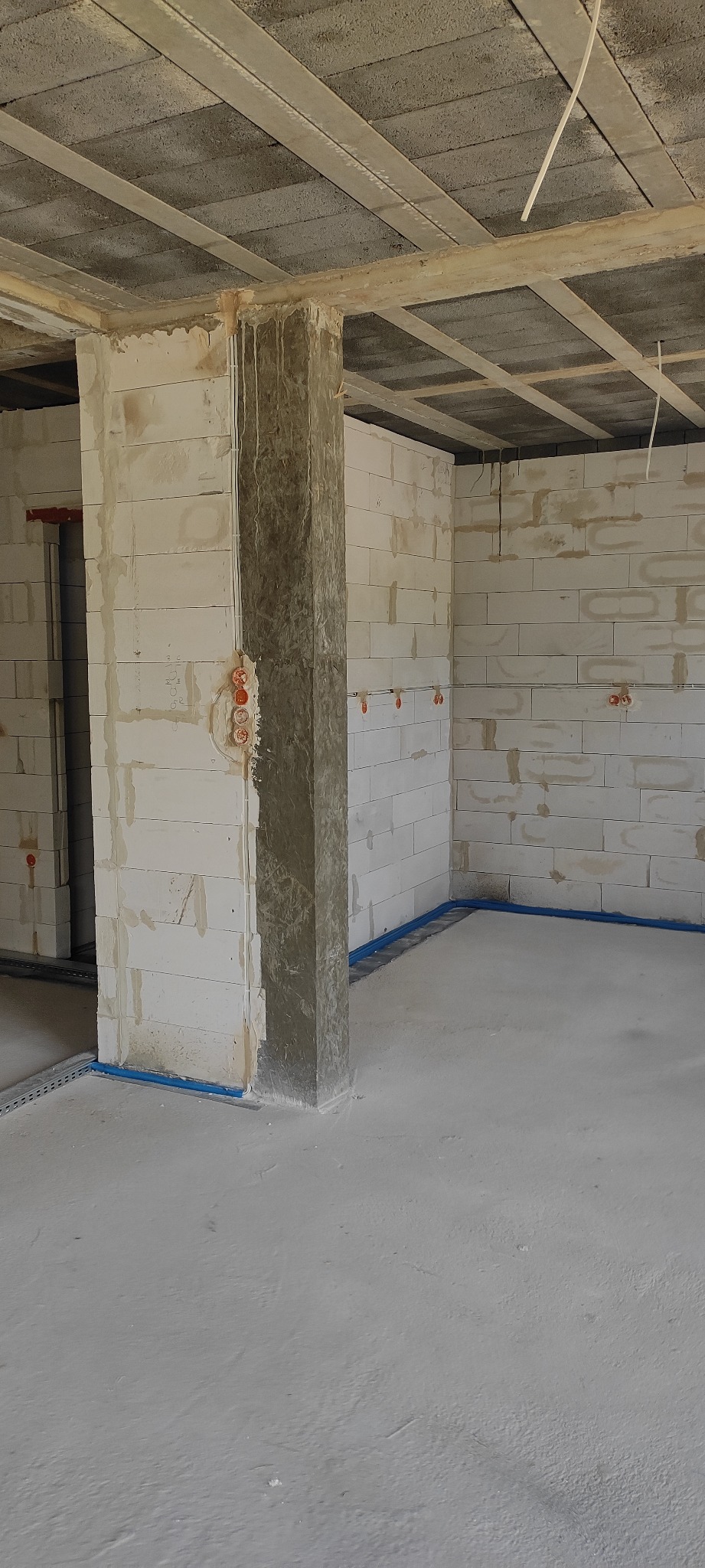 Surowy stan wnętrza budynku w trakcie budowy, widoczne ściany z betonu komórkowego, strop z betonowych belek, instalacja elektryczna w peszlach, puszki elektryczne w ścianach, niebieska rura wzdłuż...
