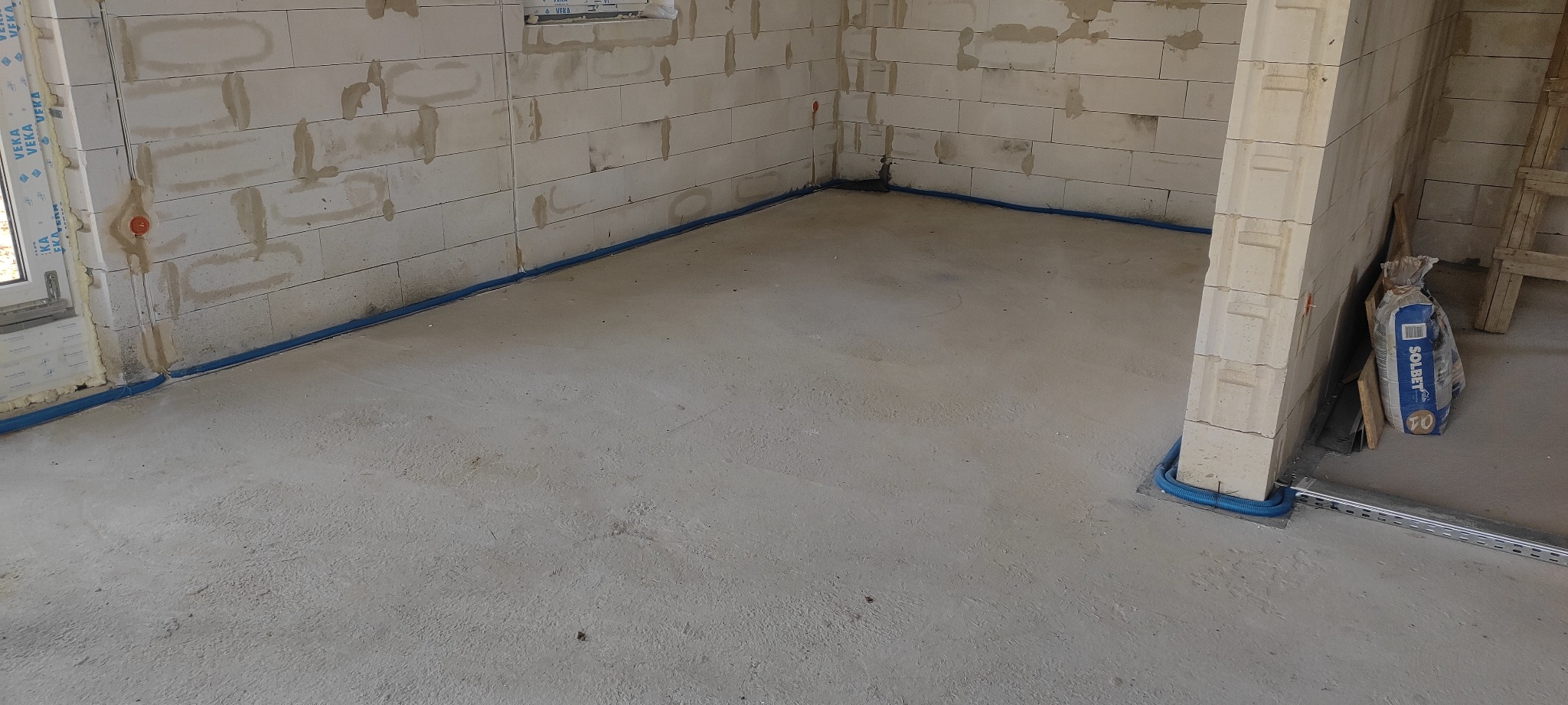 Wnętrze w trakcie budowy z widocznymi przewodami elektrycznymi w niebieskiej rurce karbowanej ułożonymi wzdłuż ściany i pod oknem, ściany z białych bloczków, w tle worek z zaprawą.