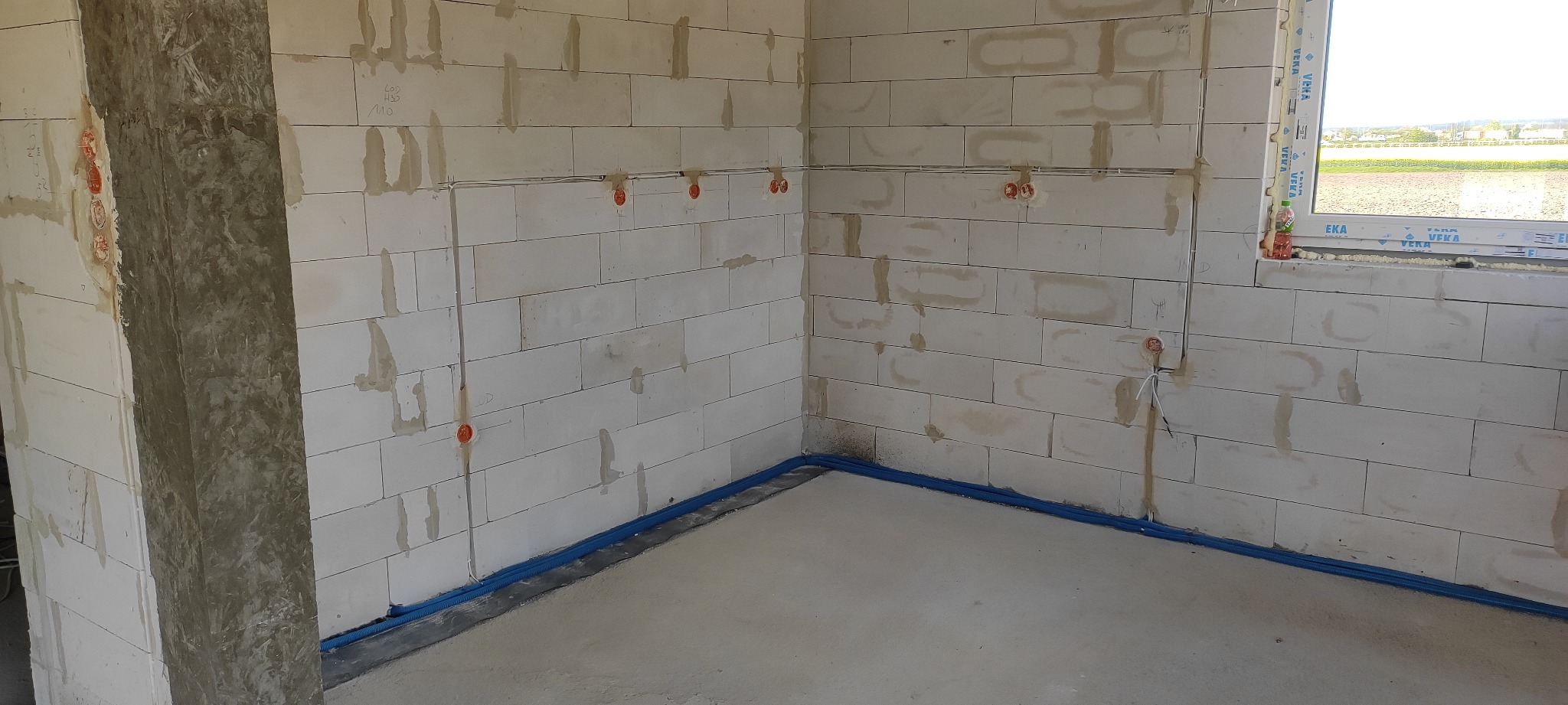 Surowy stan wnętrza z widocznymi przewodami elektrycznymi poprowadzonymi po ścianach z bloczków betonowych, zabezpieczonymi niebieską rurką przy podłodze i puszkami instalacyjnymi w ścianach.