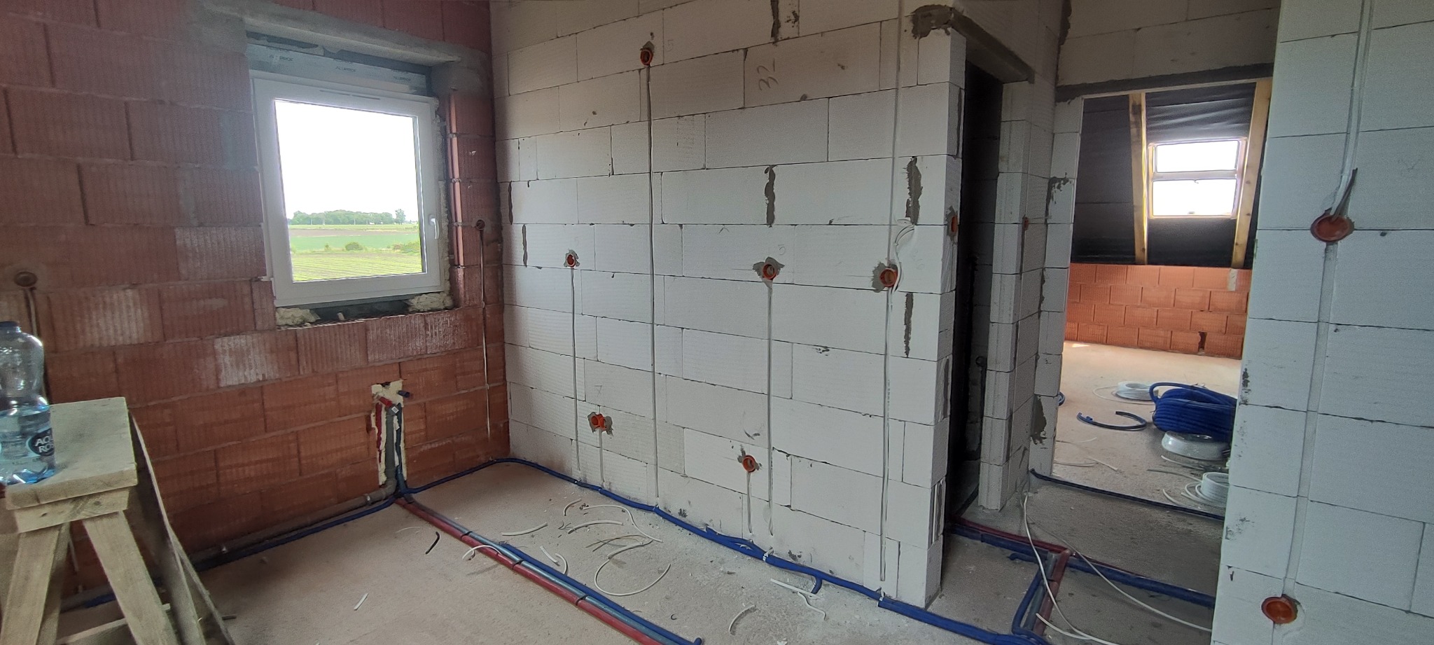 Wnętrze w stanie surowym z widocznymi przewodami elektrycznymi i rurami instalacyjnymi ułożonymi wzdłuż ścian z bloczków, puszki elektryczne w ścianach, widok przez otwór drzwiowy do kolejnego...