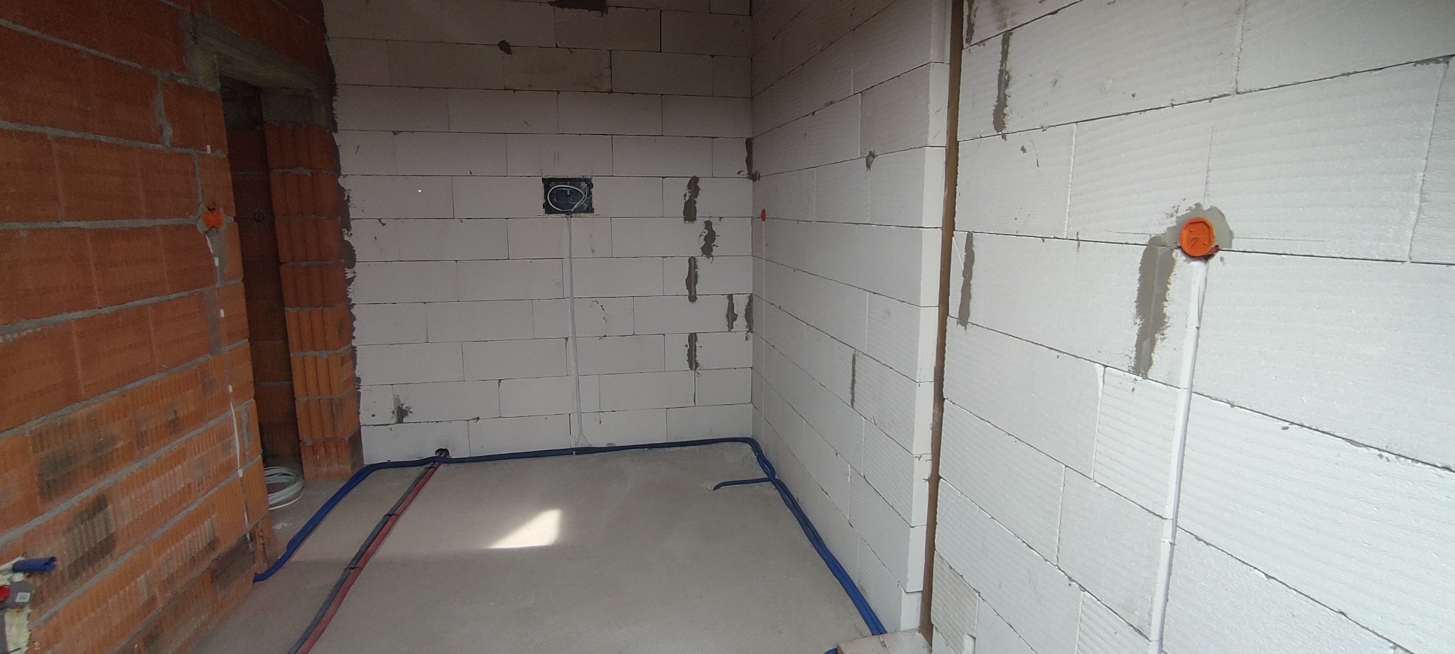 Surowy stan wnętrza w trakcie prac: ściany z białych bloczków, częściowo z czerwonej cegły, widoczne bruzdy pod kable elektryczne i rury, puszki elektryczne w ścianach, brak wykończenia.