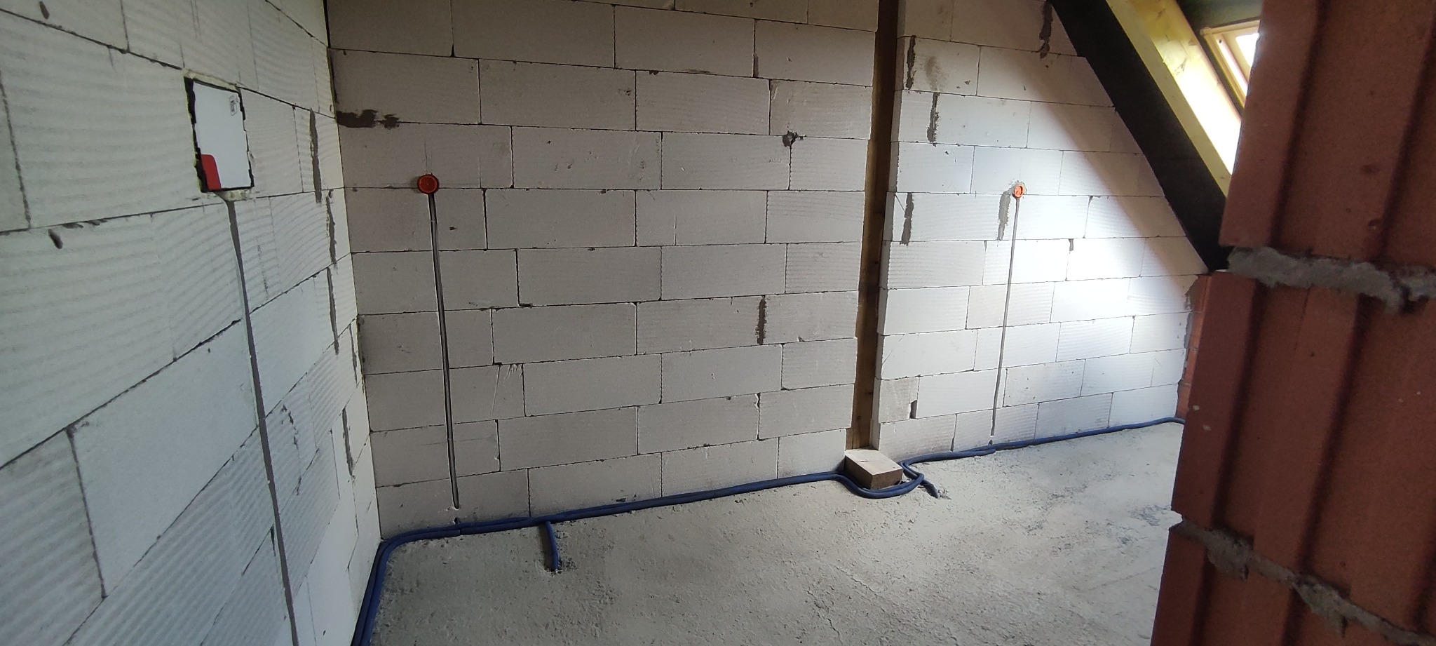 Surowy stan poddasza z widocznymi przewodami elektrycznymi w ścianach z białych bloczków, niebieska rurka instalacyjna przy podłodze i wystający fragment dachu.