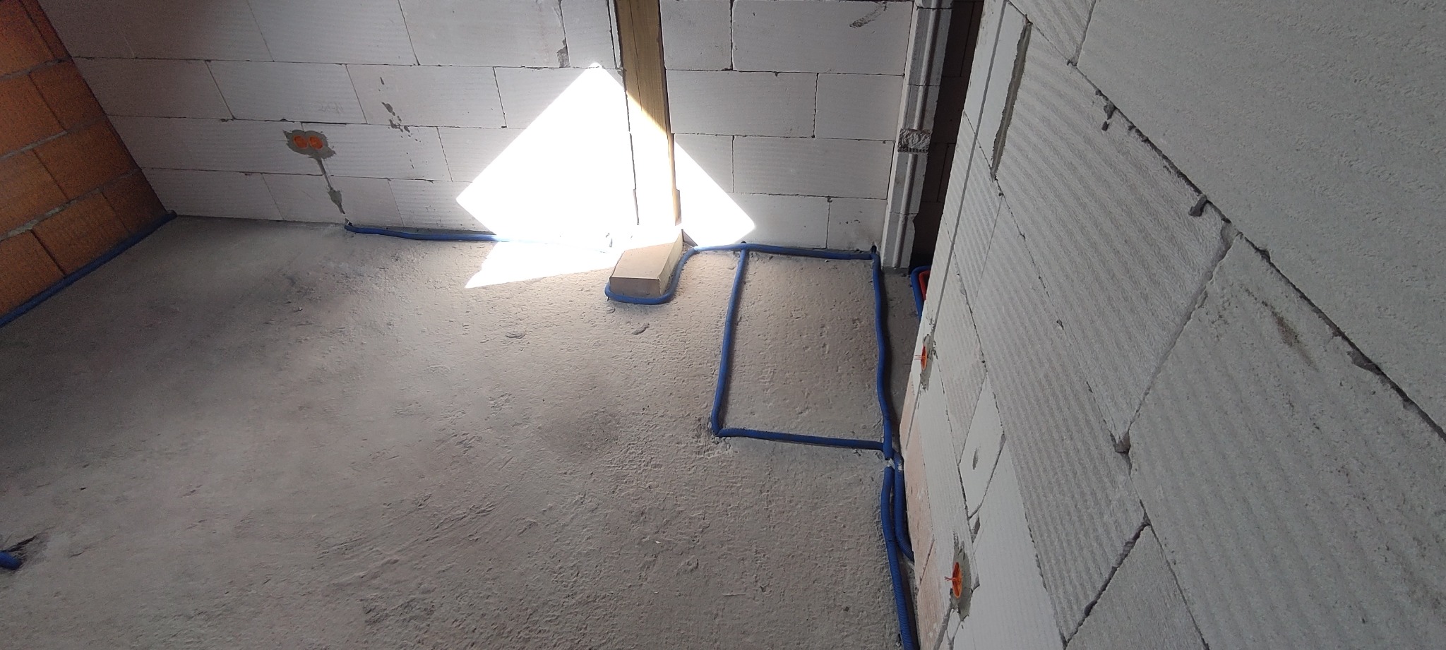 Surowy stan pomieszczenia w trakcie prac: ściany z betonu komórkowego z widocznymi puszkami elektrycznymi, niebieskie rurki instalacyjne ułożone wzdłuż ścian i podłogi, jasny promień światła...