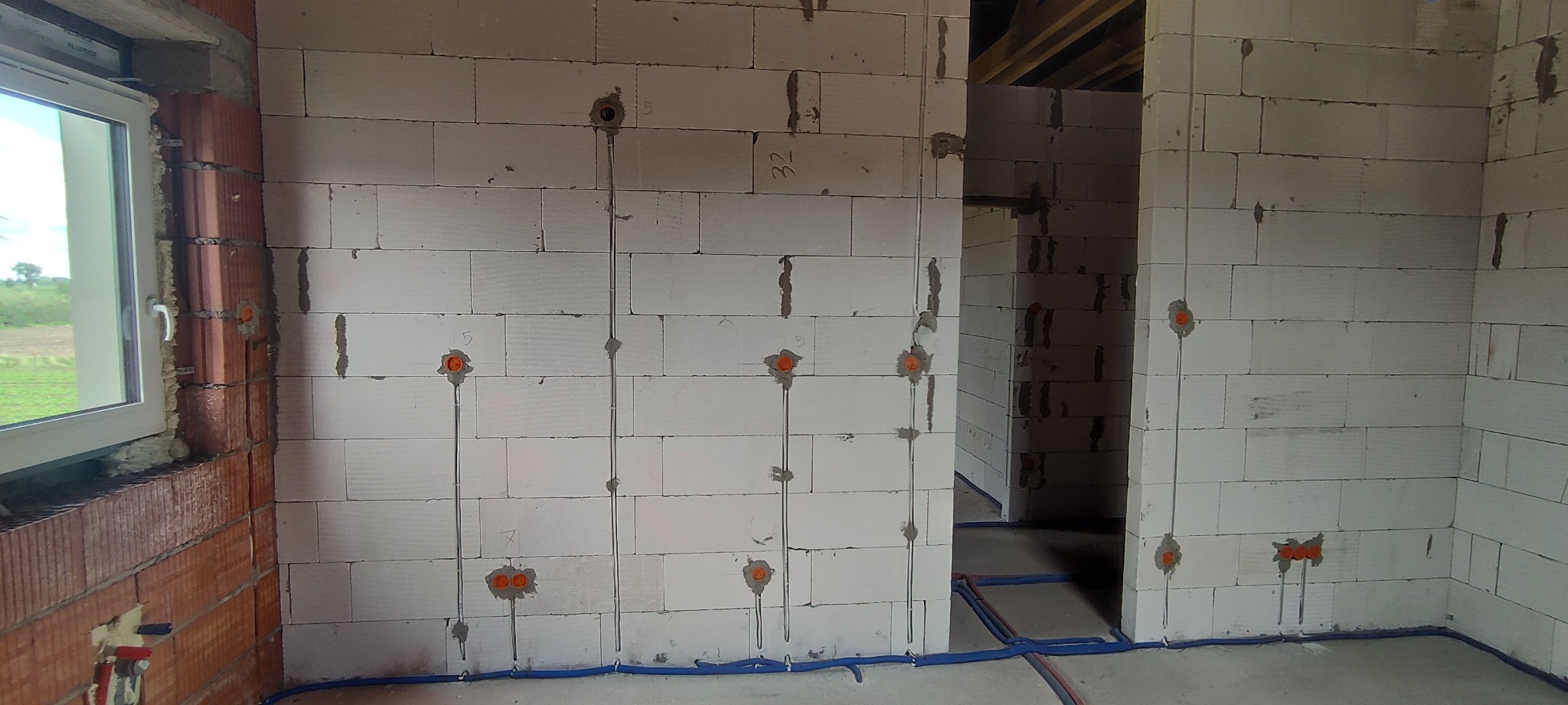 Wewnętrzne ściany z betonu komórkowego w surowym stanie, z widocznymi bruzdami pod instalację elektryczną, pomarańczowymi puszkami pod gniazdka i przewodami w podłodze, okno z widokiem na zewnątrz.