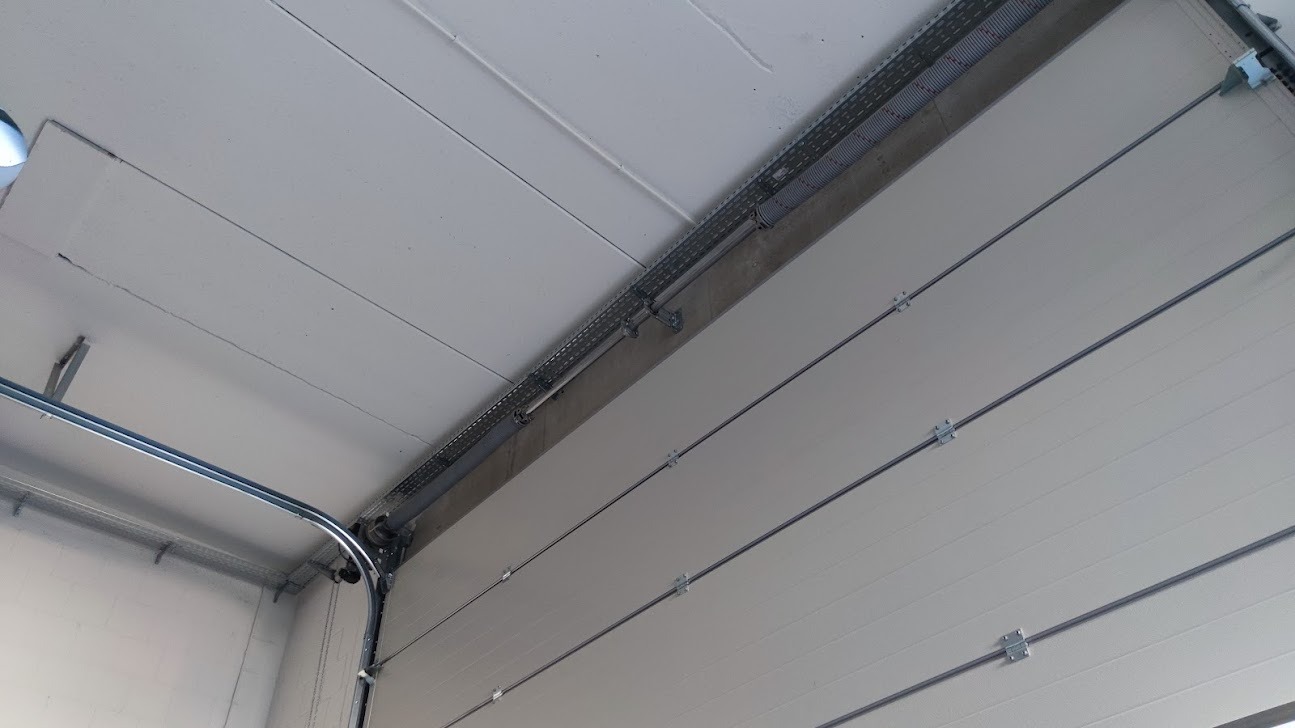Wnętrze garażu z widoczną białą segmentową bramą garażową, szynami prowadzącymi i sprężynami naciągowymi, montowanymi pod sufitem z widocznymi elementami instalacji elektrycznej w korytkach.