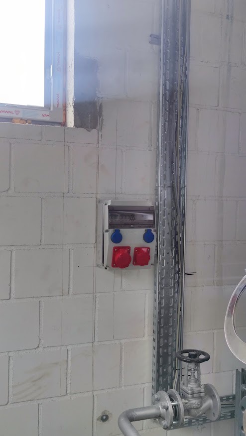 Skrzynka elektryczna z czerwonymi i niebieskimi gniazdami na ścianie z betonu komórkowego, obok pionowy kanał kablowy i fragment instalacji hydraulicznej z zaworem.