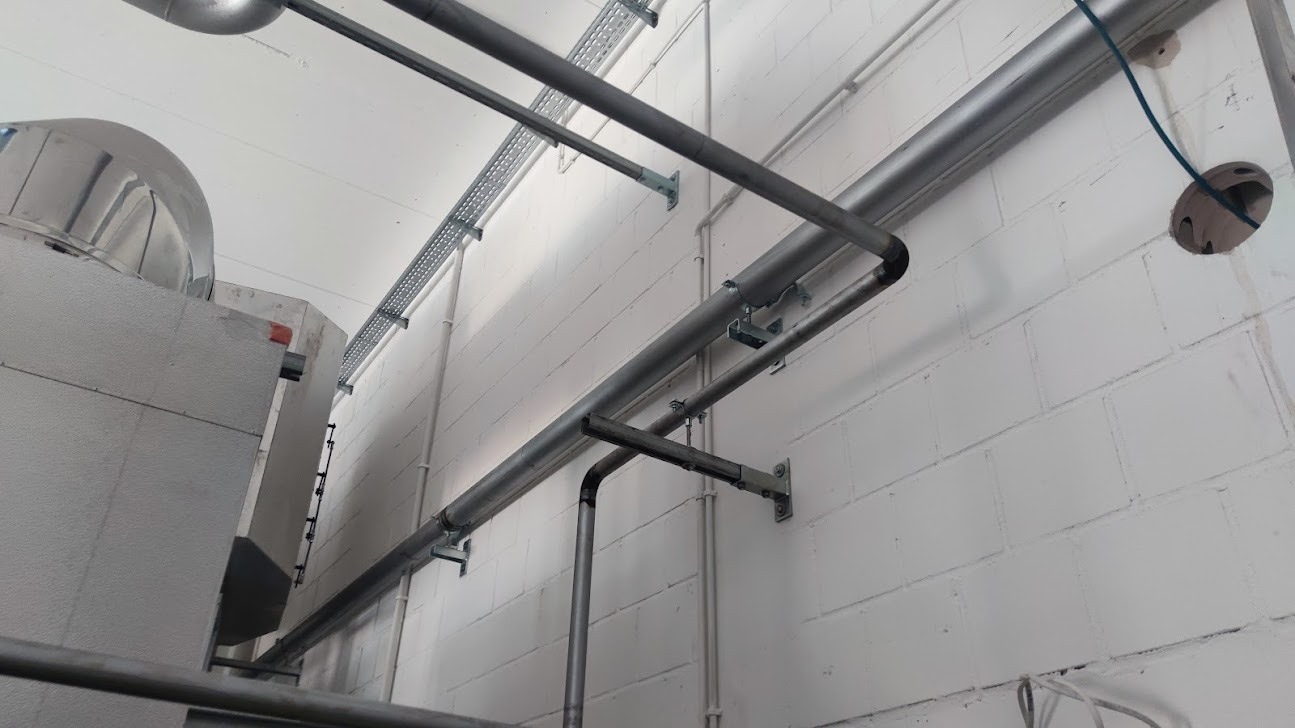 Szare rury i koryta kablowe przymocowane do białej ściany z cegieł, widoczny otwór w ścianie z wystającym kablem.