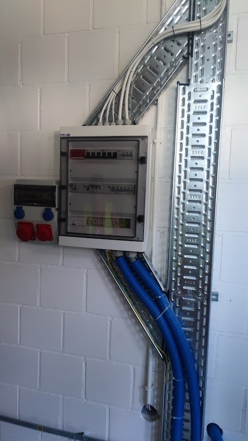 Rozdzielnica elektryczna natynkowa z zabezpieczeniami, gniazda siłowe na ścianie z bloczków, koryta kablowe z przewodami w peszlach ochronnych.