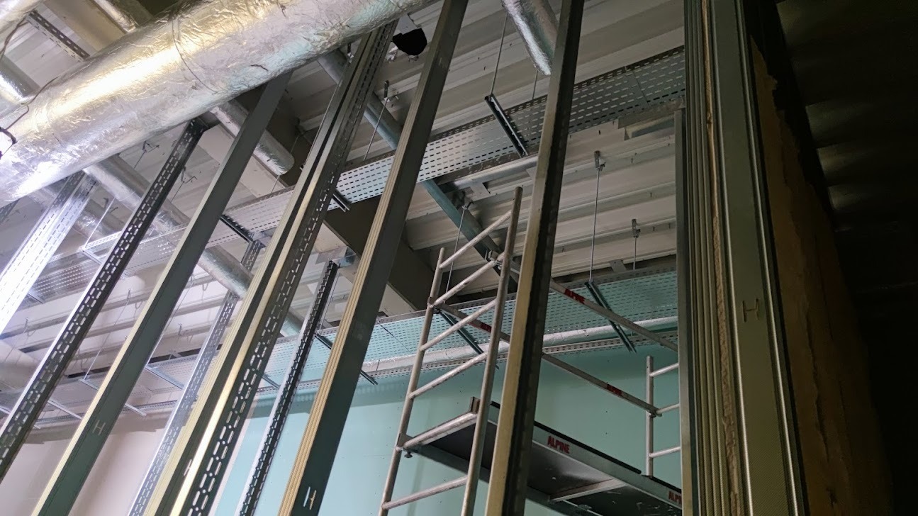 Montaż metalowych korytek kablowych pod sufitem w hali przemysłowej, widoczne rury wentylacyjne i drabina rusztowania.
