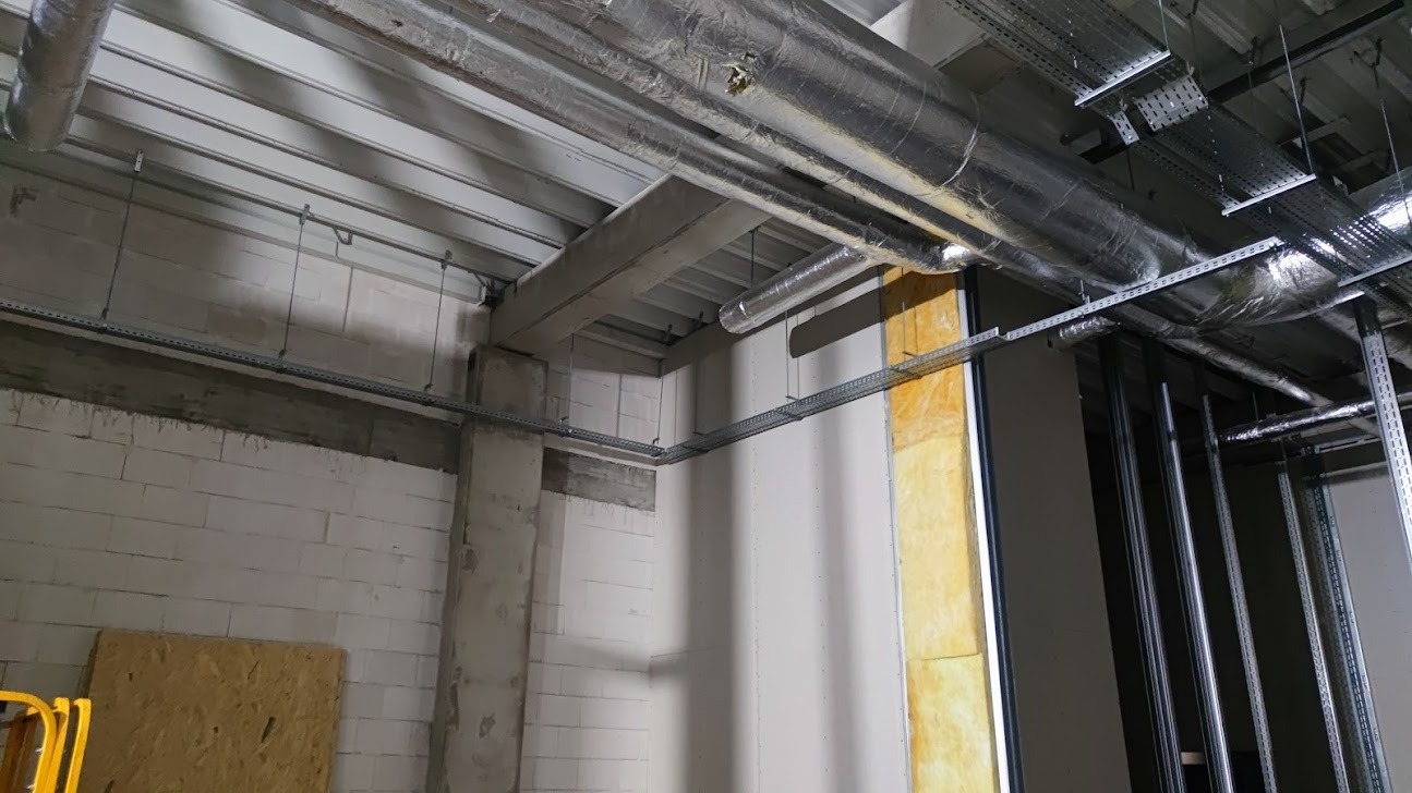 Surowy stan wnętrza w trakcie prac wykończeniowych, widoczne rury wentylacyjne z izolacją, koryta kablowe pod sufitem, ściany z betonu komórkowego i izolacja z wełny mineralnej.