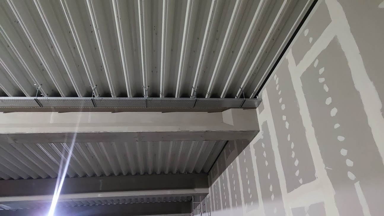 Metalowa konstrukcja sufitowa z perforowanymi profilami montażowymi i zawiesiami, widoczna na tle surowej ściany z płyt gipsowo-kartonowych z widocznymi łączeniami.