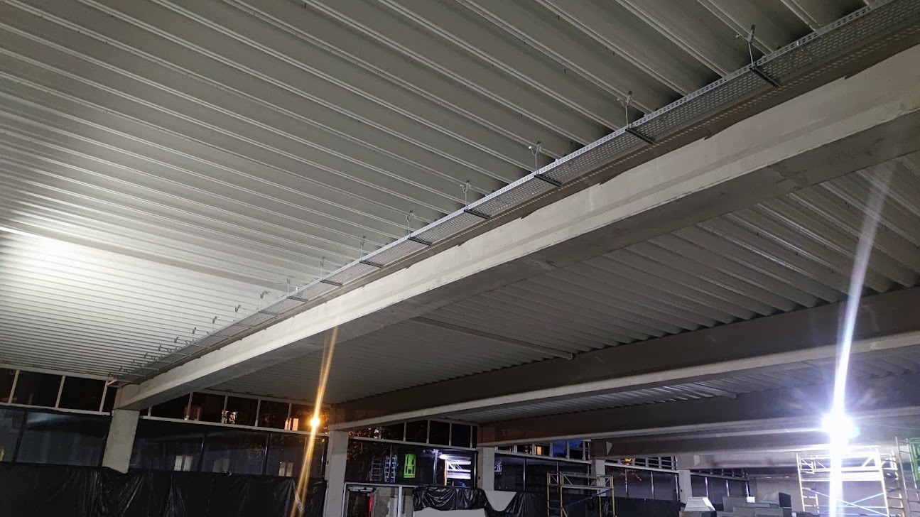 Metalowa konstrukcja sufitu z zamontowanymi korytkami kablowymi, widoczne elementy konstrukcyjne budynku i fragmenty otoczenia zewnętrznego przez okna.