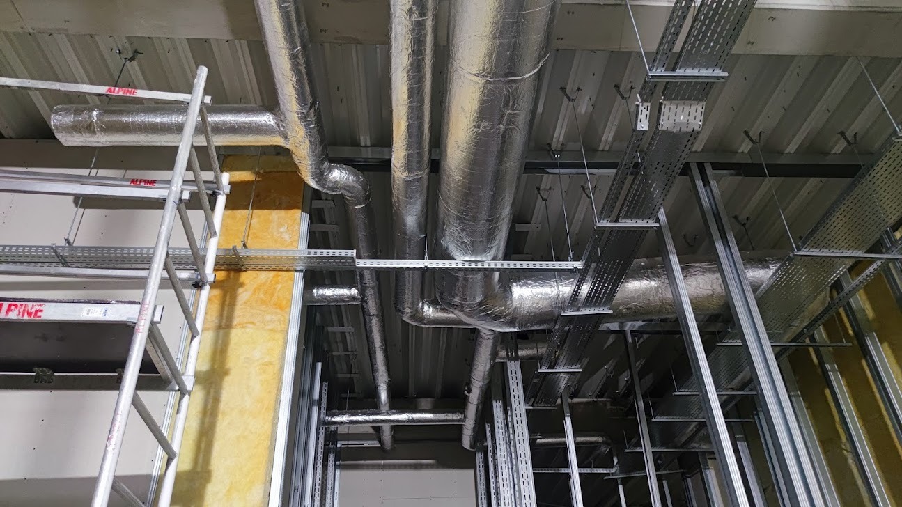 Widok sufitu w trakcie montażu instalacji wentylacyjnych i tras kablowych, widoczne ocieplone rury wentylacyjne, metalowe koryta kablowe oraz fragment rusztowania.