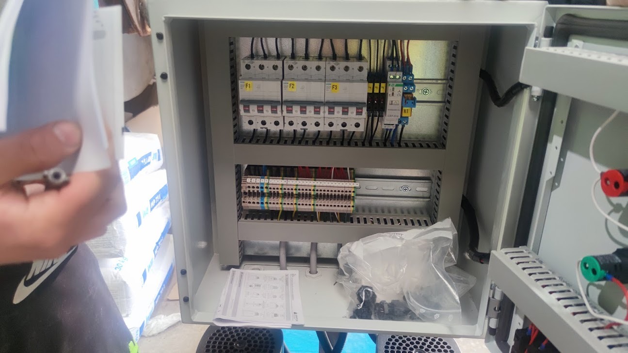 Otwarta szafa sterownicza z widocznymi bezpiecznikami, listwami zaciskowymi i przewodami, częściowo zakryta przez dłoń trzymającą dokumentację techniczną, w tle worki z materiałami budowlanymi.