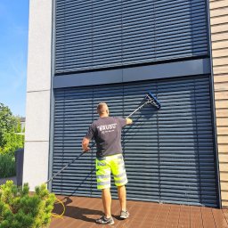 CPB CZYŚCIOCH POGROMCA BRUDU - Pracownik w neonowych szortach czyści długą szczotką żaluzje fasadowe nowoczesnego budynku, stojąc na drewnianym tarasie.