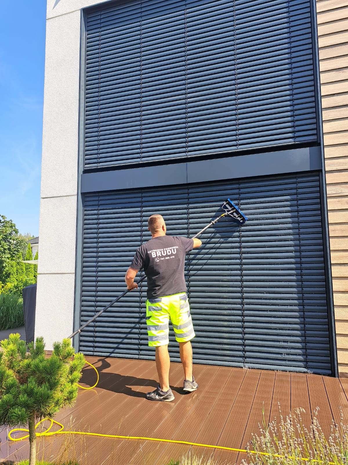 Pracownik w neonowych szortach czyści długą szczotką żaluzje fasadowe nowoczesnego budynku, stojąc na drewnianym tarasie.