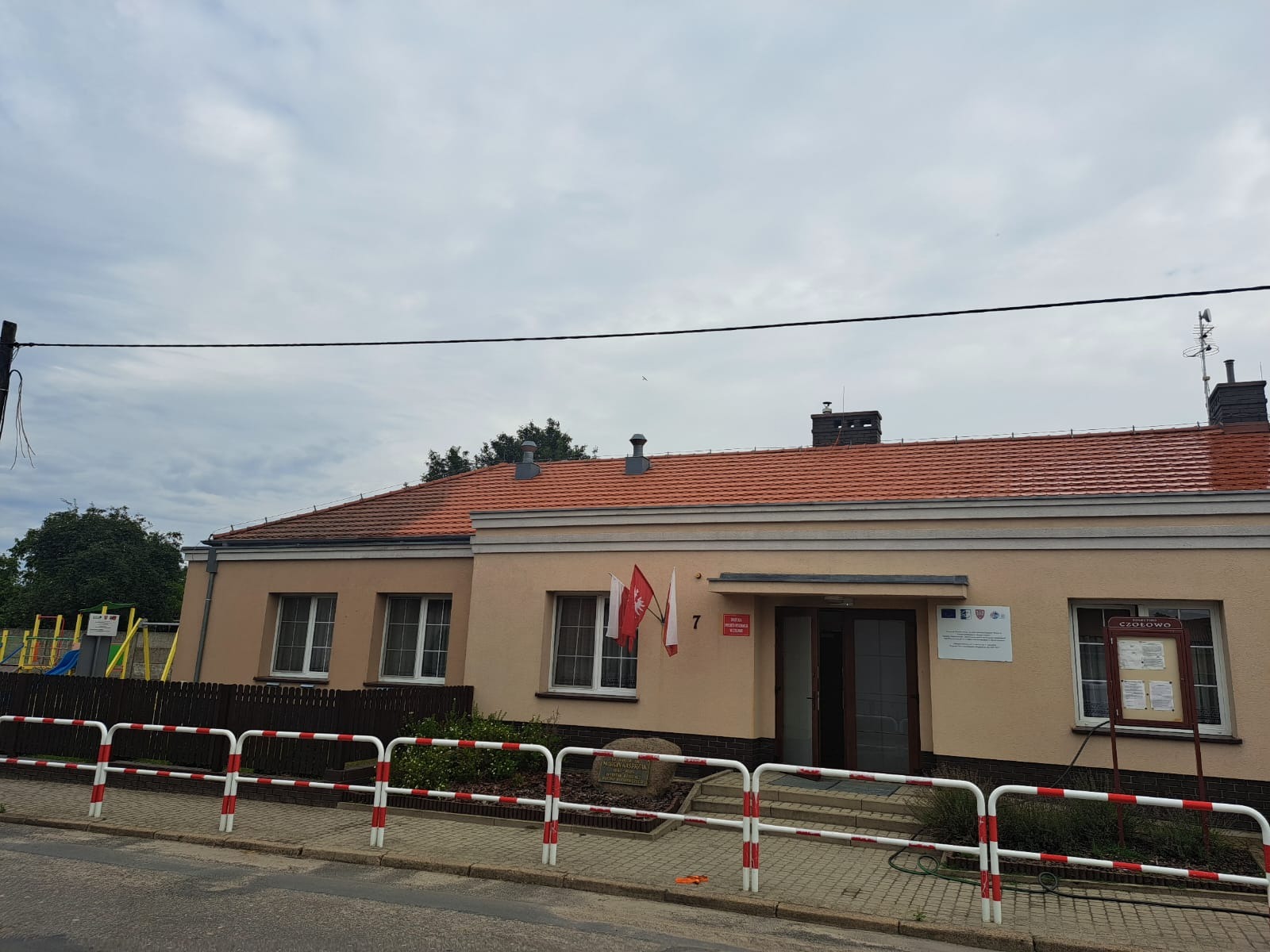 Budynek parterowy z pomarańczową dachówką, otynkowany na beżowo, z dwoma polskimi flagami i tablicami informacyjnymi przy wejściu, otoczony niskim, czerwono-białym płotkiem i chodnikiem z kostki.