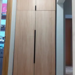 Wysoka szafa wnękowa z jasnego drewna z długimi, czarnymi uchwytami, umieszczona w wąskim korytarzu z widocznymi drzwiami po obu stronach.