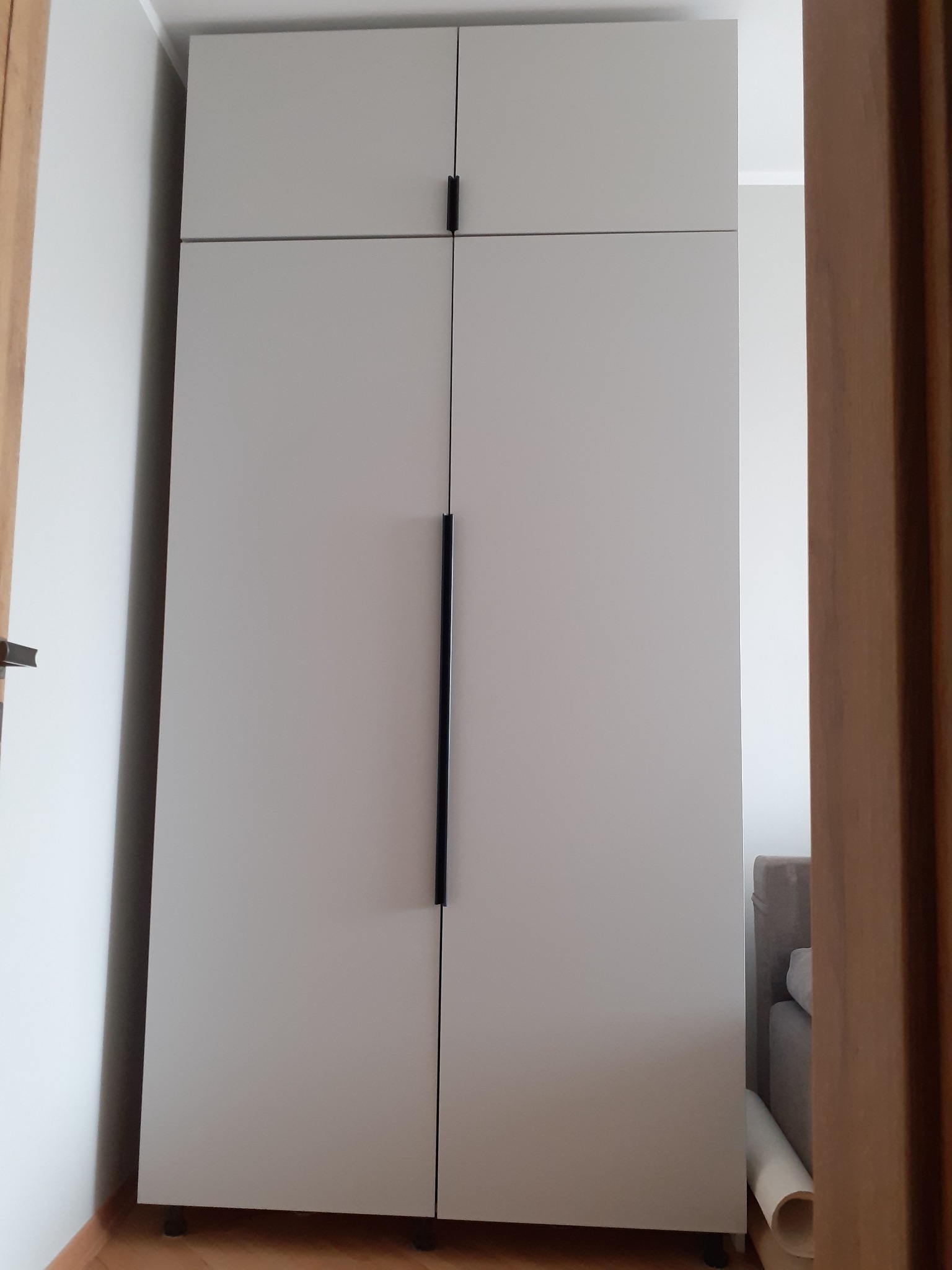 Jasnoszara szafa dwudrzwiowa z czarnymi, minimalistycznymi uchwytami, widoczna z perspektywy wejścia do pokoju, z fragmentem łóżka w tle.