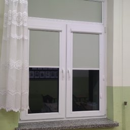 F.H.U. Artur Grabarczyk - Okna w klasie w Węgrowie z częściowo zasłoniętymi roletami wewnętrznymi w kolorze jasnoszarym oraz białą, koronkową firanką po lewej stronie.