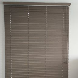 F.H.U. Artur Grabarczyk - Zamknięte, poziome, brązowe listwy w oknie, z delikatnym prześwitem światła, sznurek do regulacji zwisa po prawej stronie, na suficie widoczny wystający kabel.