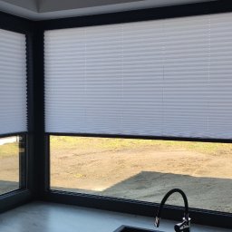 F.H.U. Artur Grabarczyk - Białe, plisowane osłony okienne zamontowane w narożnym oknie z czarną ramą, widok na zewnątrz na teren z ziemią i trawą, fragment blatu kuchennego z baterią w dolnej części kadru.