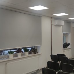 F.H.U. Artur Grabarczyk - Szare rolety wewnętrzne w sali konferencyjnej z klimatyzacją i roślinami na parapetach, widok na projektor sufitowy i flagi w tle.