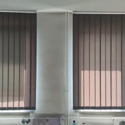 Dwa okna z szarymi pionowymi panelami osłaniającymi przed światłem, widoczne prążki światła słonecznego, na parapecie suszące się pranie i ręczniki.