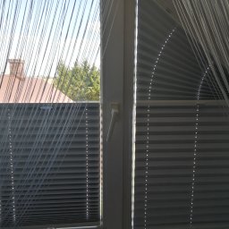 F.H.U. Artur Grabarczyk - Okno z zamontowanymi plisami w kolorze szarym, z widokiem na dach domu i drzewa, zasłonięte dodatkowo dekoracyjną zasłoną sznurkową.