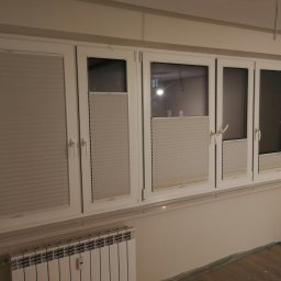 F.H.U. Artur Grabarczyk - Szereg białych okien z zamontowanymi jasnoszarymi plisami, niektóre z nich częściowo zasłonięte, widoczny grzejnik pod oknem i folia ochronna na podłodze.