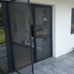 F.H.U. Artur Grabarczyk - Otwarta moskitiera ramkowa w kolorze antracytowym, zamontowana w drzwiach tarasowych, widok z zewnątrz na taras z betonową posadzką i wnętrze domu.