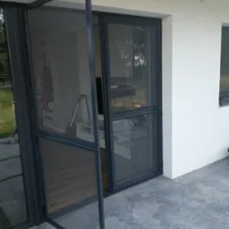 Otwarta moskitiera ramkowa w kolorze antracytowym, zamontowana w drzwiach tarasowych, widok z zewnątrz na taras z betonową posadzką i wnętrze domu.