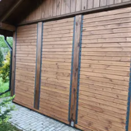 Drewniana zabudowa balkonu z przesuwnymi panelami, widok z zewnątrz, zieleń w tle. Naturalne oświetlenie podkreśla strukturę drewna.