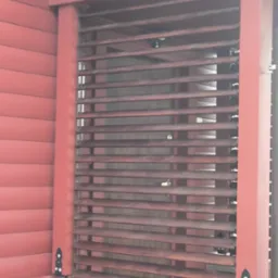 Czerwona, pozioma przesłona okienna z drewna, zamontowana na tle ściany z desek w tym samym kolorze. Widoczne detale mocowania i konstrukcji.