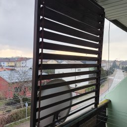 F.H.U. Artur Grabarczyk - Drewniane żaluzje balkonowe w kolorze ciemnego brązu, zamontowane na balkonie z widokiem na ulicę i zabudowę miejską w pochmurny dzień.