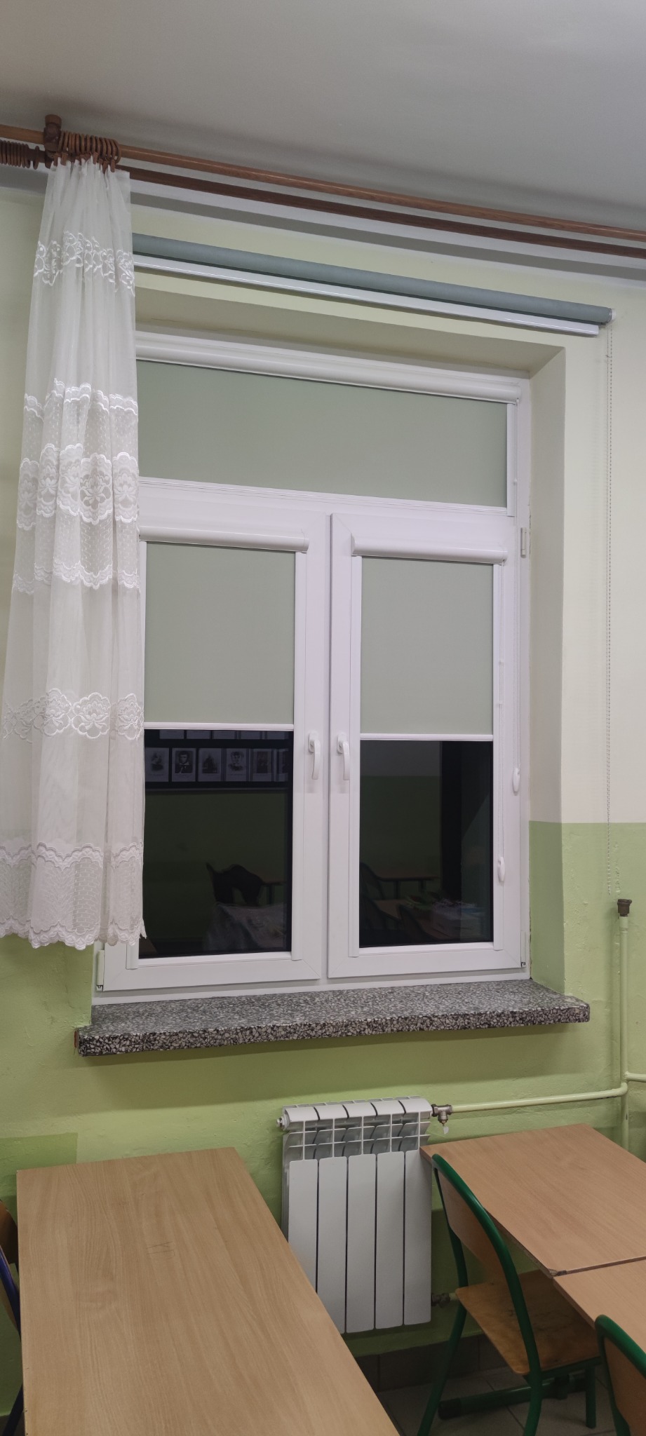 Okna w klasie w Węgrowie z częściowo zasłoniętymi roletami wewnętrznymi w kolorze jasnoszarym oraz białą, koronkową firanką po lewej stronie.