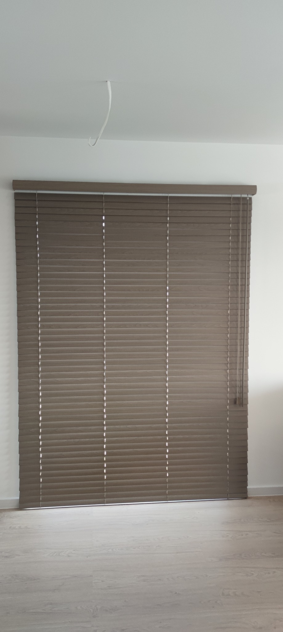 Zamknięte, poziome, brązowe listwy w oknie, z delikatnym prześwitem światła, sznurek do regulacji zwisa po prawej stronie, na suficie widoczny wystający kabel.