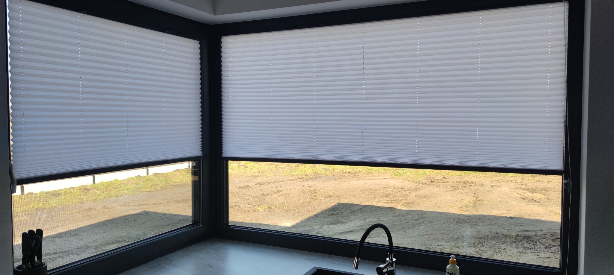 Białe, plisowane osłony okienne zamontowane w narożnym oknie z czarną ramą, widok na zewnątrz na teren z ziemią i trawą, fragment blatu kuchennego z baterią w dolnej części kadru.