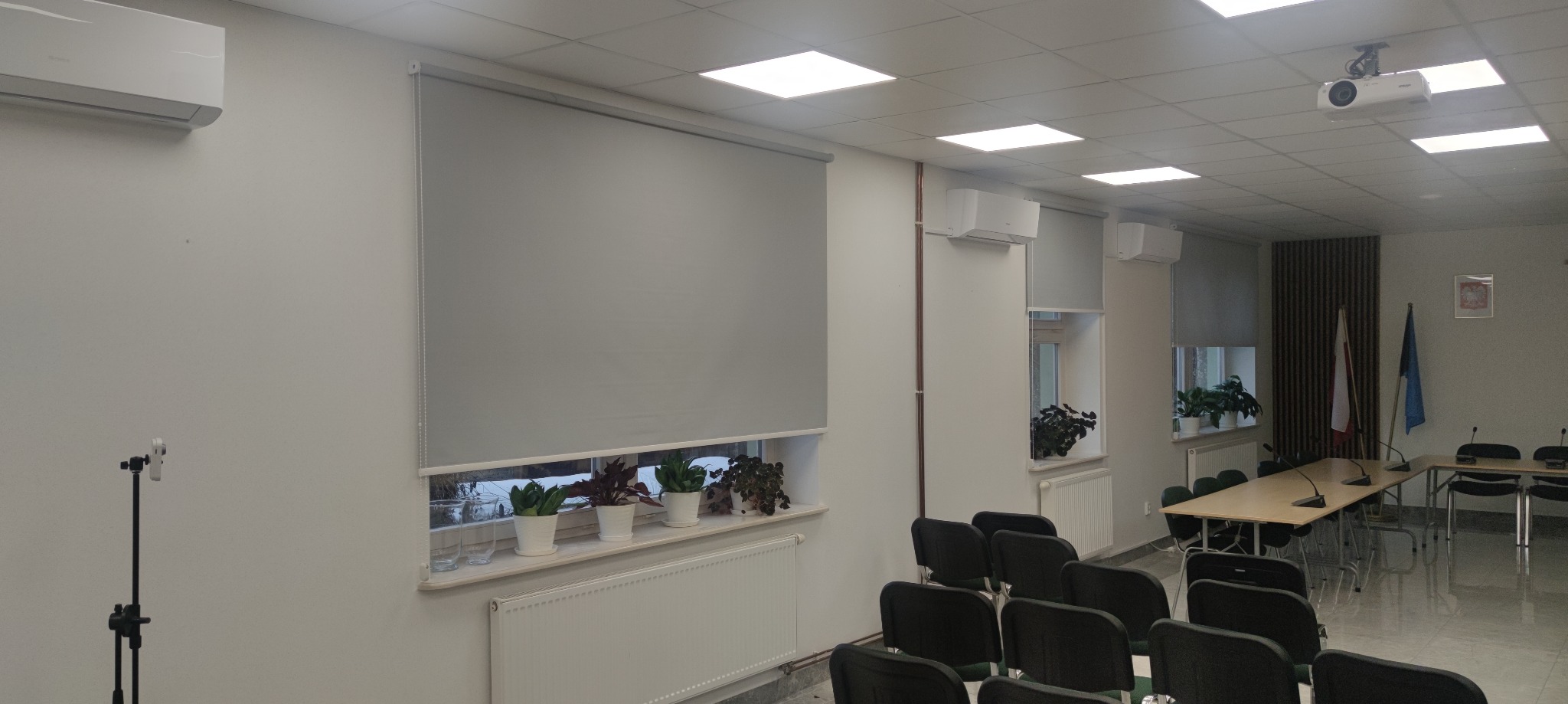 Szare rolety wewnętrzne w sali konferencyjnej z klimatyzacją i roślinami na parapetach, widok na projektor sufitowy i flagi w tle.