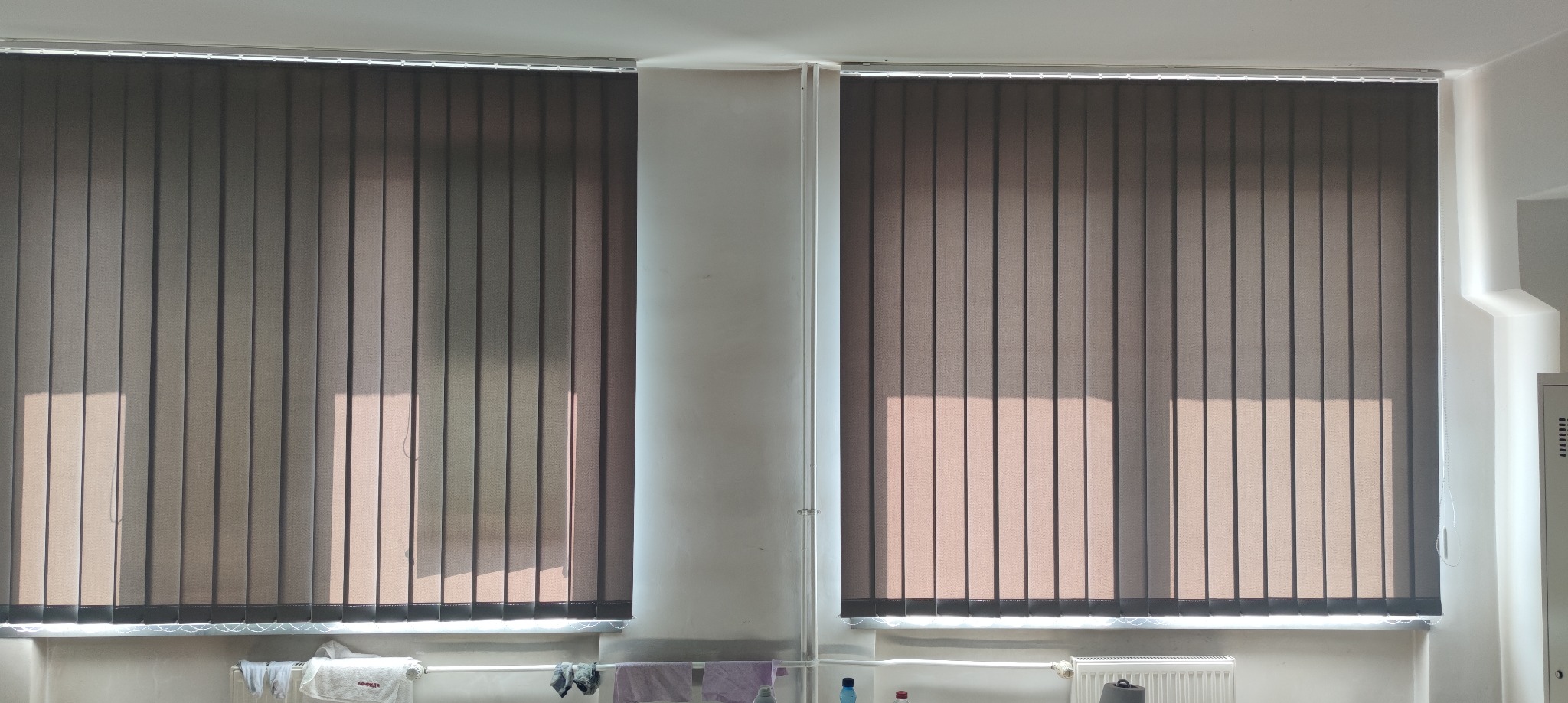 Dwa okna z szarymi pionowymi panelami osłaniającymi przed światłem, widoczne prążki światła słonecznego, na parapecie suszące się pranie i ręczniki.